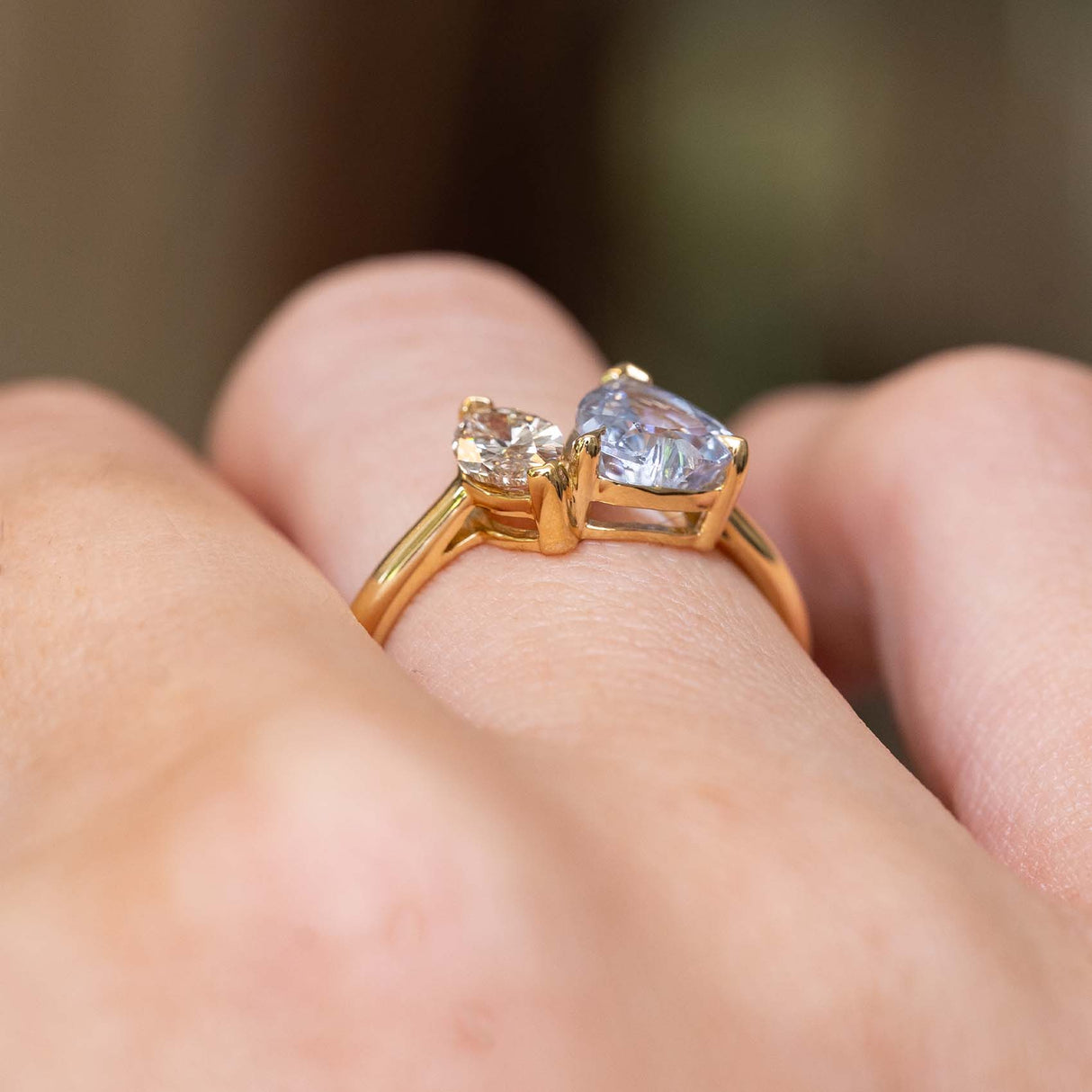 A close-up photograph of a hand wearing an antique-inspired, two stone toi et moi yellow gold engagement ring of a large colourless sapphire and a brown diamond seen from the side to showcase the open gallery and claw setting-style.