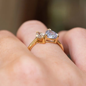 A close-up photograph of a hand wearing an antique-inspired, two stone toi et moi yellow gold engagement ring of a large colourless sapphire and a brown diamond seen from the side to showcase the open gallery and claw setting-style.