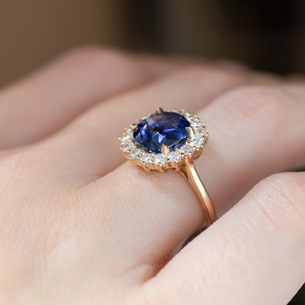 Angled close-up of the blue sapphire cluster engagement ring worn on a finger, showing the depth of the gemstone and its surrounding diamonds.