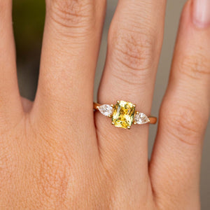 A close-up of a hand wearing an Antique-inspired, emerald-cut yellow sapphire and diamond three-stone ring against a blurred background.