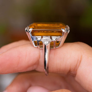 A close-up of an Antique, Art Deco citrine and diamond cocktail ring in platinum worn on a person's finger, seen from the side to show off the gallery
