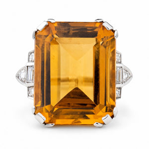 An antique emerald-cut orange citrine cocktail ring set in platinum flanked by diamonds on each side shown on a plain white background.