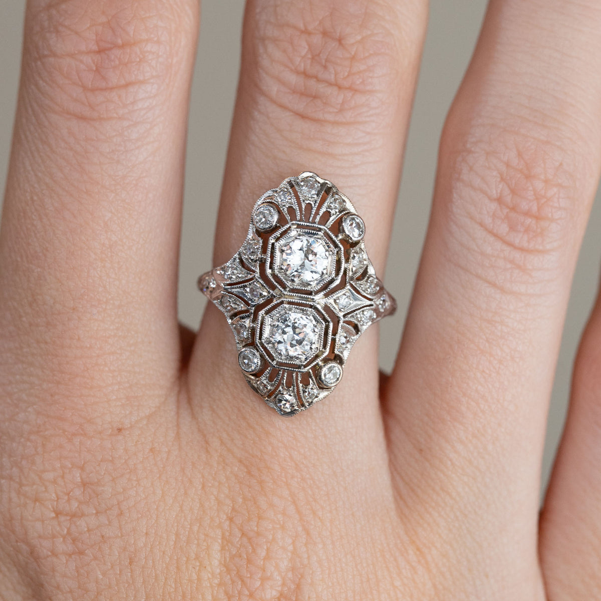 Close-up of the antique platinum ring on a finger, displaying two prominent diamonds framed by intricate geometric patterns and smaller accent stones.