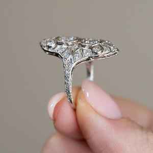 Side profile of the platinum filigree ring held between fingers, showcasing its elevated diamond setting, intricate gallery and slender engraved band.