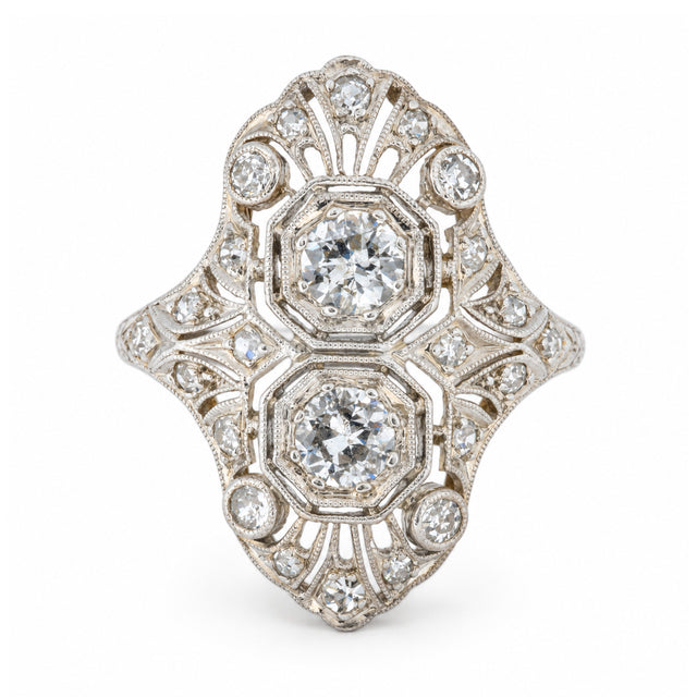 An antique Art Deco platinum cocktail ring featuring two central diamonds within an elaborate geometric filigree design, accented by smaller diamonds.