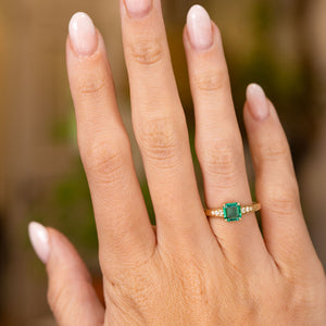  An outstretched hand wearing a yellow gold engagement ring with an emerald flanked by small diamonds shown to display the proportions