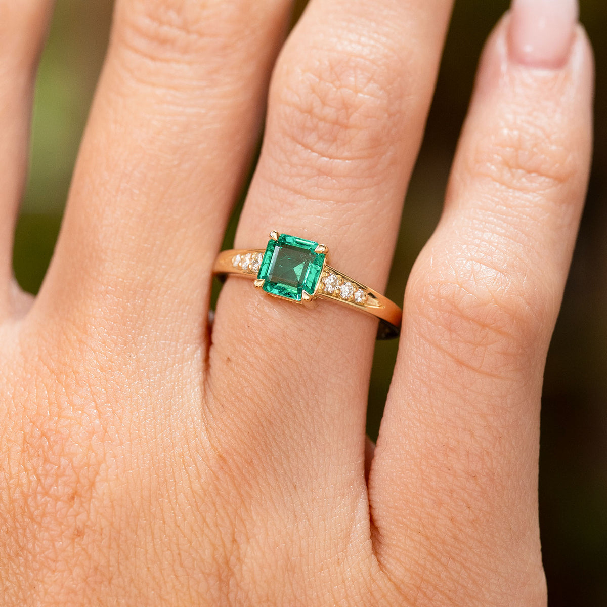 A close-up of a hand wearing a Vintage-inspired, Emerald and diamond flanked solitaire engagement ring.