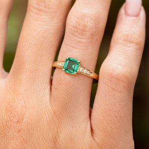 A close-up of a hand wearing a Vintage-inspired, Emerald and diamond flanked solitaire engagement ring.