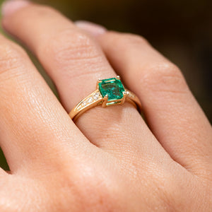 A Vintage-inspired, Emerald and diamond flanked solitaire engagement ring in yellow gold with an open gallery worn on the finger and seen from a low angle to show the setting style.