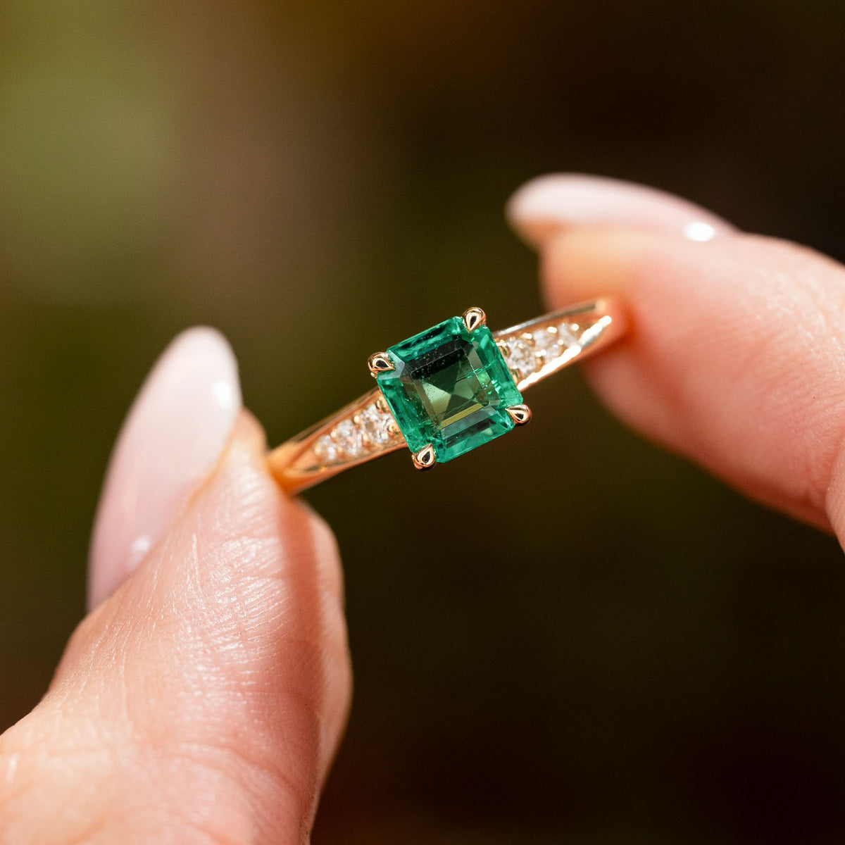 A vintage inspired yellow gold engagement ring with an emerald and diamond set shoulders held in between two fingers and shown up close.