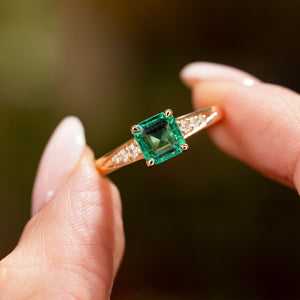 A vintage inspired yellow gold engagement ring with an emerald and diamond set shoulders held in between two fingers and shown up close.