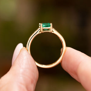 A Vintage-inspired, Emerald and diamond flanked solitaire engagement ring in yellow gold held up in profile view, shoving the setting style and shank.