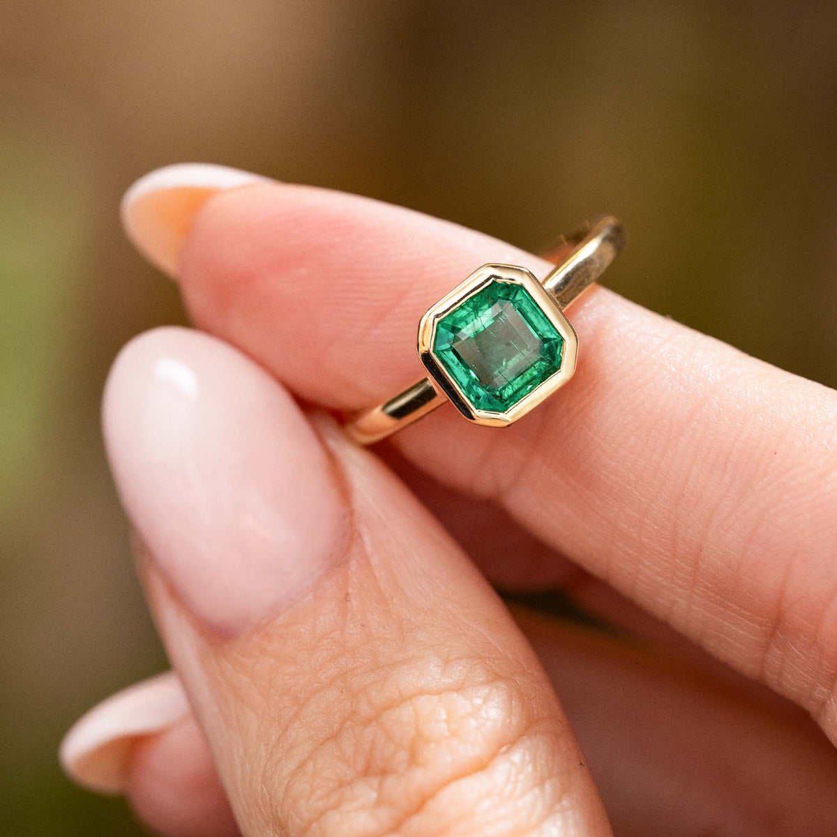 A close-up of a hand holding a yellow gold Antique-inspired, Emerald solitaire engagement ring set against a soft, blurred natural background.