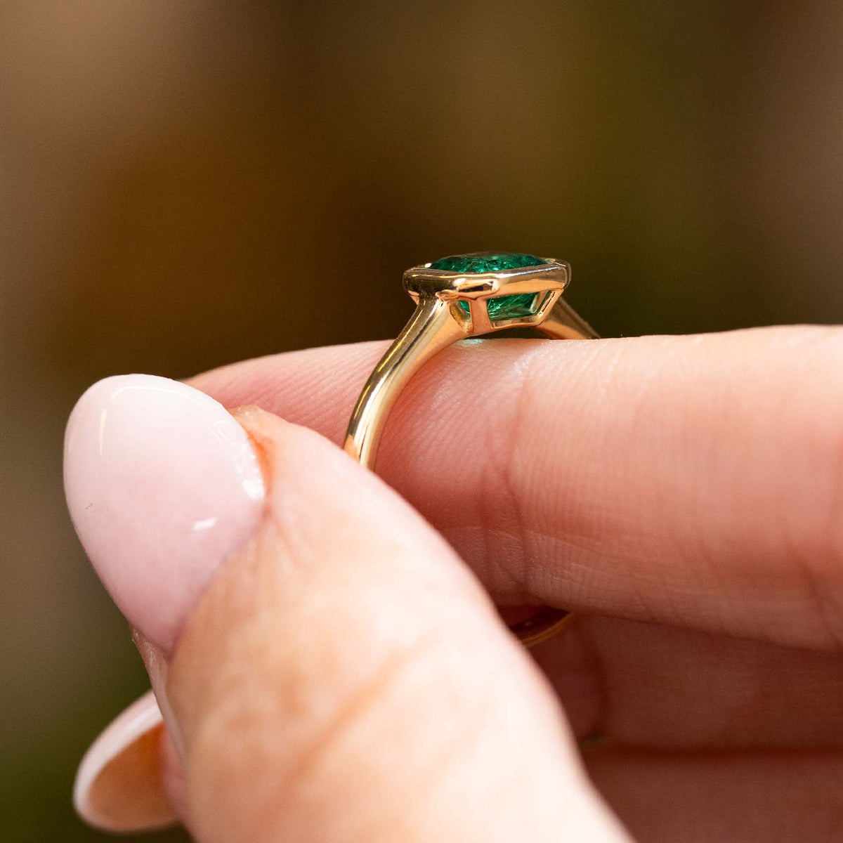 A close-up of a gold ring with a green rectangular gemstone being held between fingers, the ring, Antique-inspired, Emerald solitaire engagement ring.