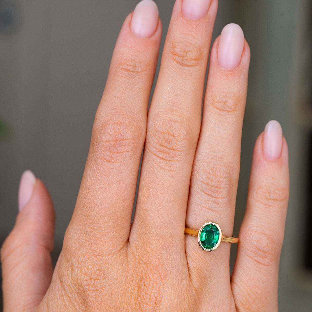 A close-up of a hand wearing an Antique inspired gold solitaire engagement ring featuring an oval-cut emerald, set against a soft, blurred gray background.