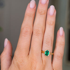 A close-up of a hand wearing an Antique inspired gold solitaire engagement ring featuring an oval-cut emerald, set against a soft, blurred gray background.