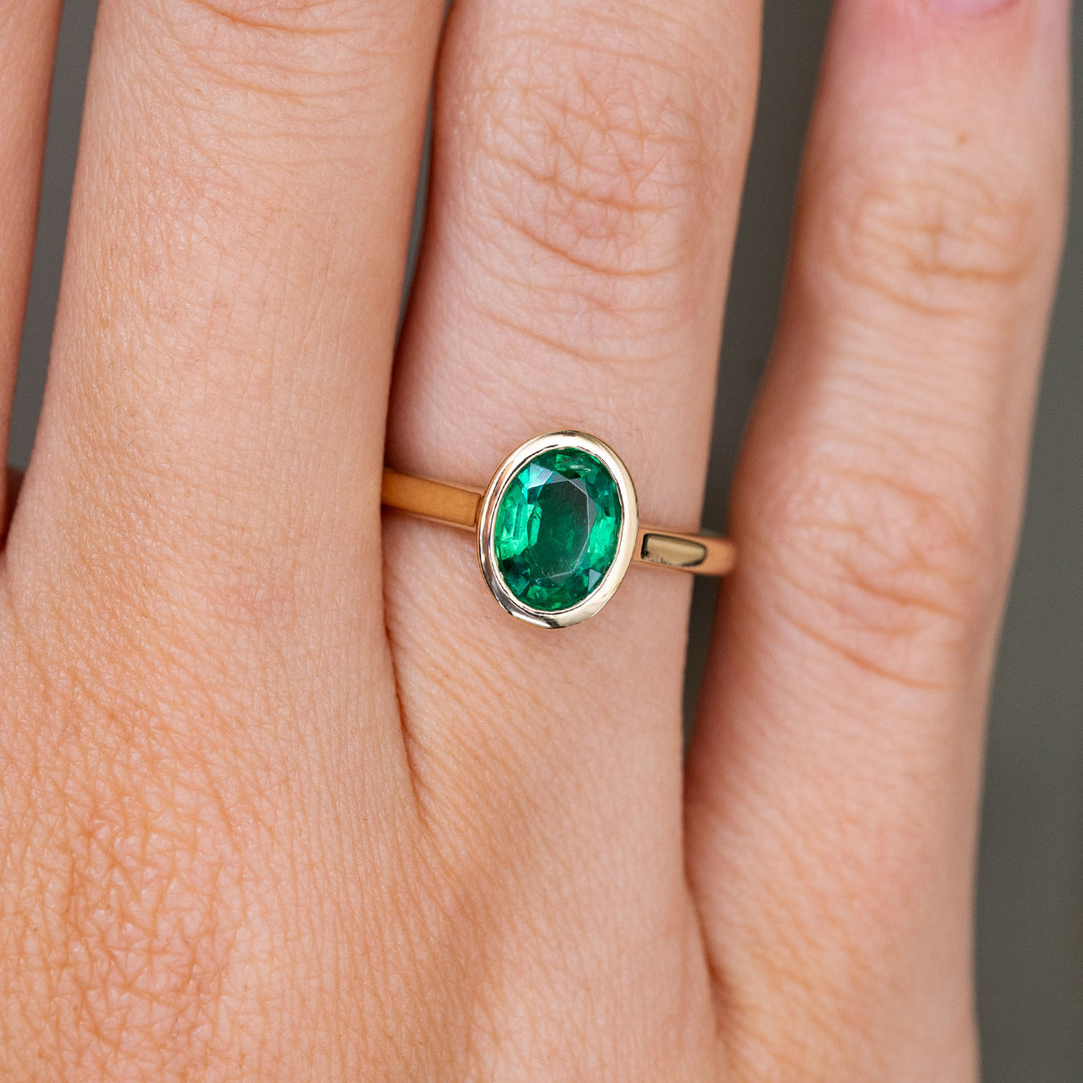 A close up of an oval emerald set in a Vintage inspired solitaire engagement ring, showcasing the yellow gold bezel setting and band worn on a finger. 