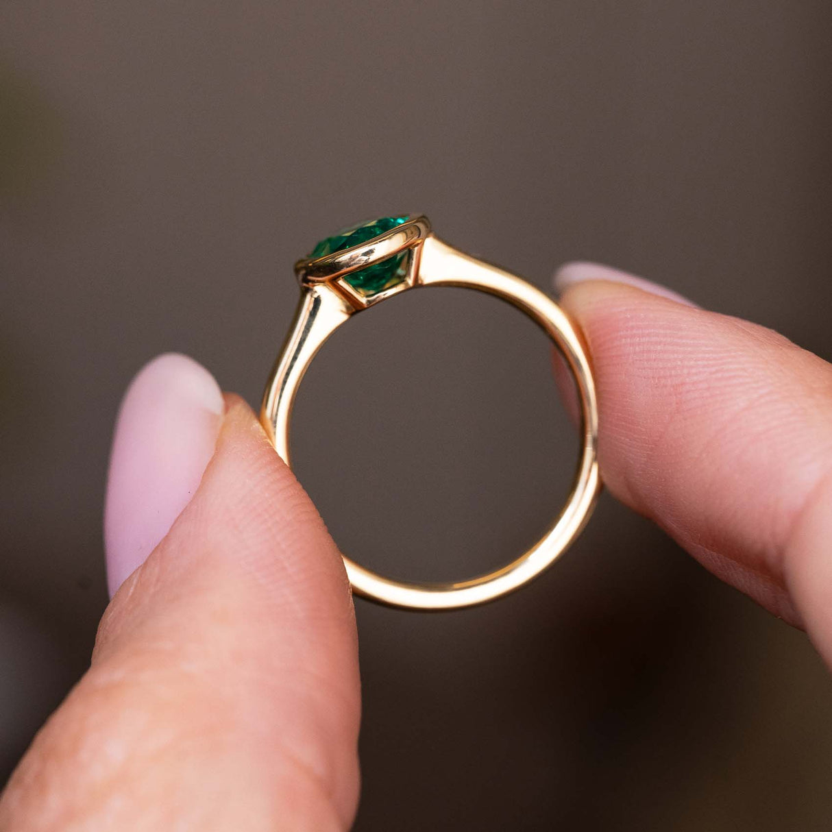 A close-up of the side profile and yellow gold setting and band of an Antique-inspired, Emerald solitaire engagement ring held between two fingers against a soft, blurred brown background.