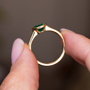 A close-up of the side profile and yellow gold setting and band of an Antique-inspired, Emerald solitaire engagement ring held between two fingers against a soft, blurred brown background.