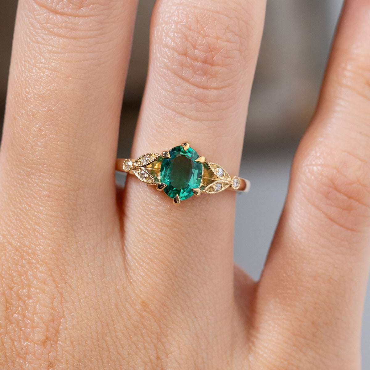 Close-up of a vintage-inspired emerald and diamond engagement ring worn on a finger, featuring a vivid green oval emerald framed by decorative yellow gold laurel leaf-inspired shoulders.