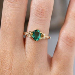 Close-up of a vintage-inspired emerald and diamond engagement ring worn on a finger, featuring a vivid green oval emerald framed by decorative yellow gold laurel leaf-inspired shoulders.