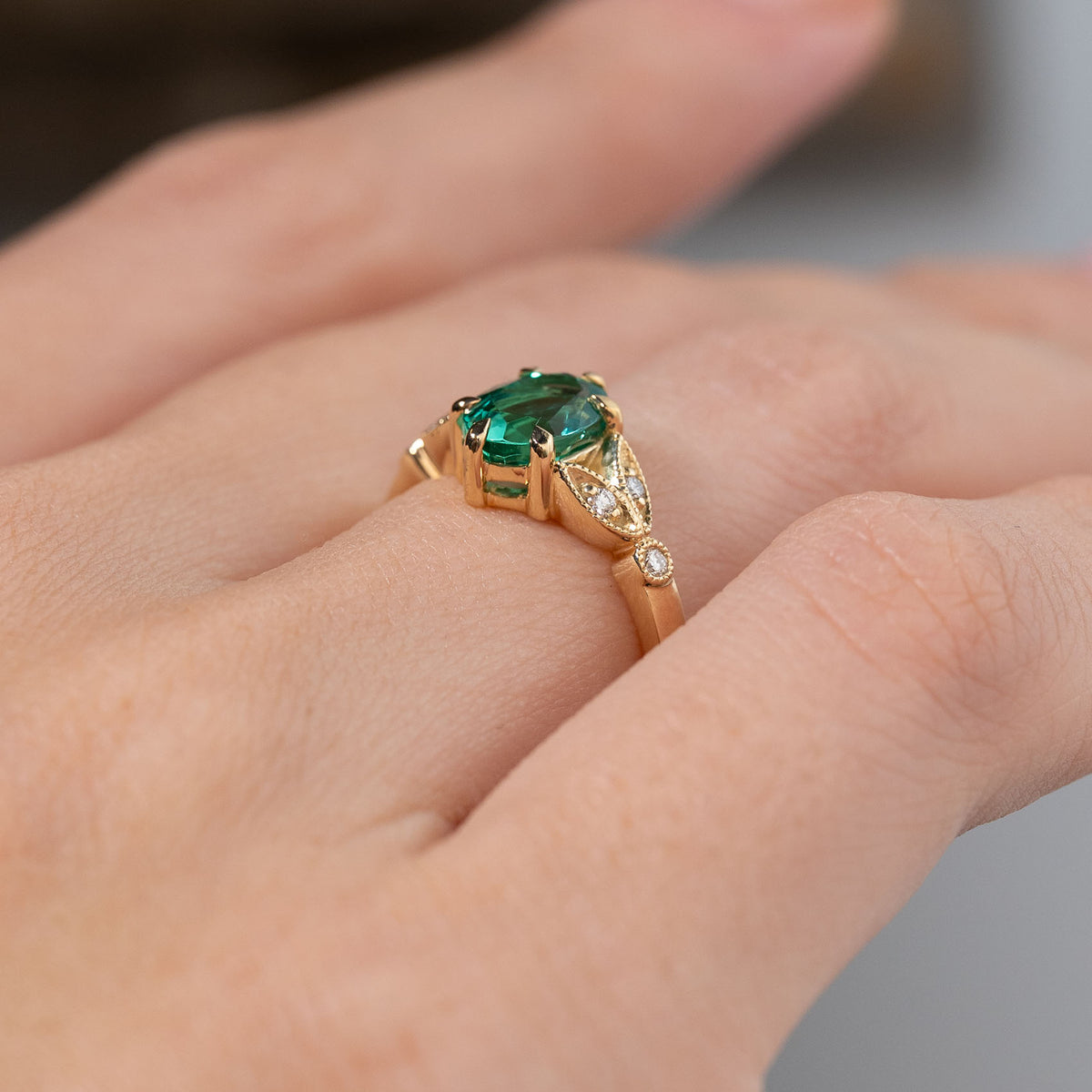 Side profile of a vintage-inspired emerald engagement ring worn on a finger, revealing its raised claw setting, decorative trefoil shoulders and polished yellow gold band.