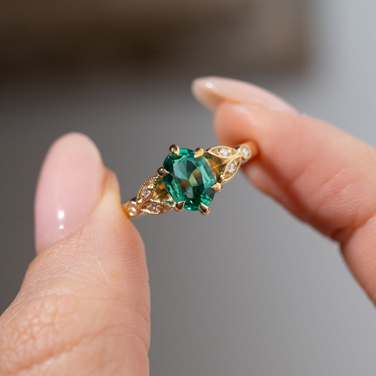 Detailed close-up of a vintage-inspired emerald and diamond engagement ring held between fingers, highlighting the faceted green emerald and ornate diamond-set laurel leaf-inspired shoulders.