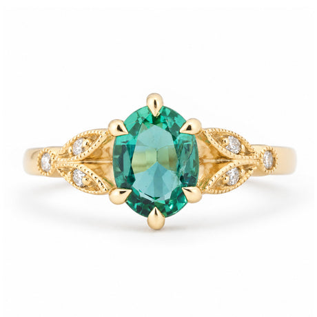 Vintage-inspired emerald and diamond engagement ring in yellow gold, featuring an oval-cut green emerald with intricate laurel leaf-inspired trefoil shoulders and diamond accents.