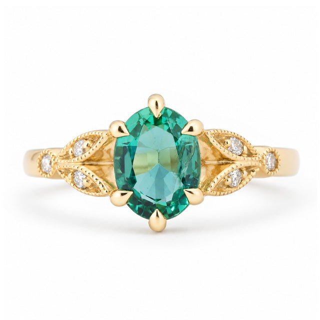 Vintage-inspired emerald and diamond engagement ring in yellow gold, featuring an oval-cut green emerald with intricate laurel leaf-inspired trefoil shoulders and diamond accents.