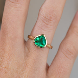 Close finger detail highlighting the bright green emerald and warm yellow gold bezel setting.