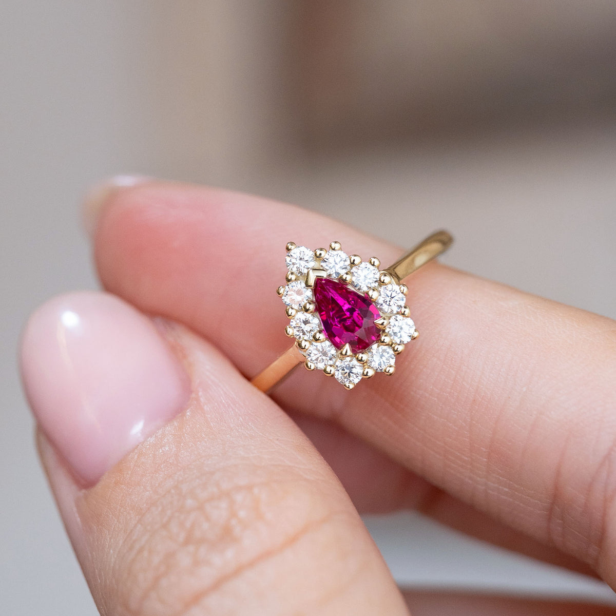 Pear-cut ruby engagement ring held between the fingers, revealing the bright diamond halo and gold setting.