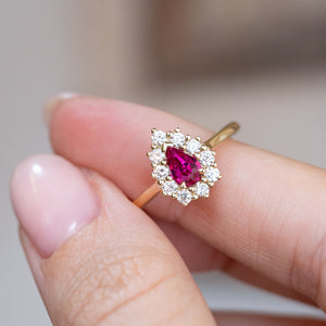 Pear-cut ruby engagement ring held between the fingers, revealing the bright diamond halo and gold setting.