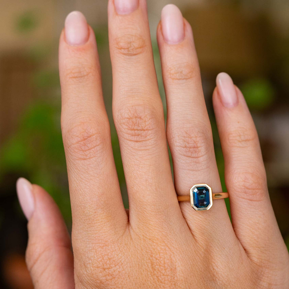 A close-up of a hand wearing an Antique-inspired, Deep marine-blue sapphire solitaire engagement ring with a yellow gold band, set against a softly blurred green background.