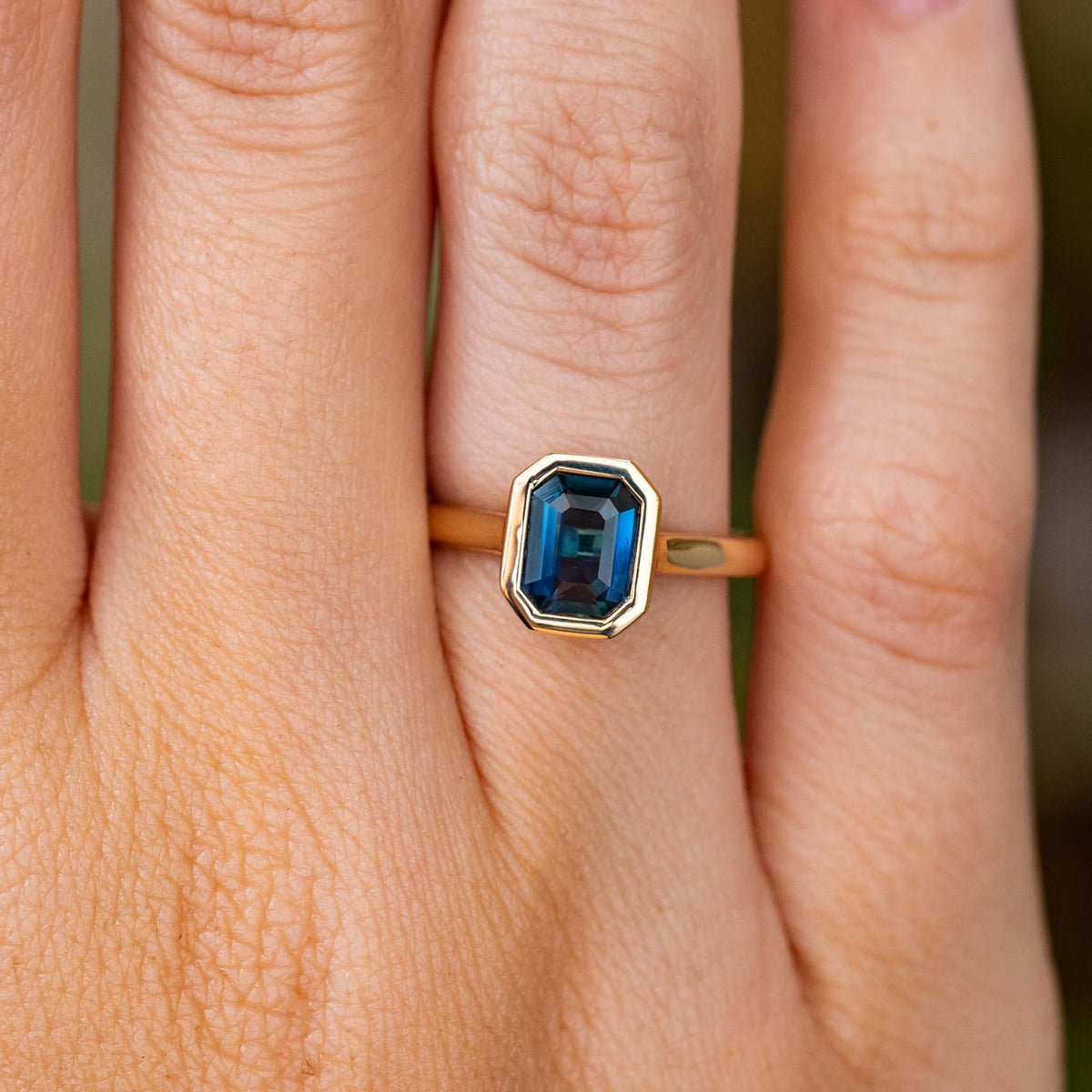 A close-up of the emerald-cut deep blue Sri Lankan sapphire set in a yellow gold Antique inspired solitaire engagement ring, worn on a finger.