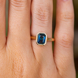 A close-up of the emerald-cut deep blue Sri Lankan sapphire set in a yellow gold Antique inspired solitaire engagement ring, worn on a finger.