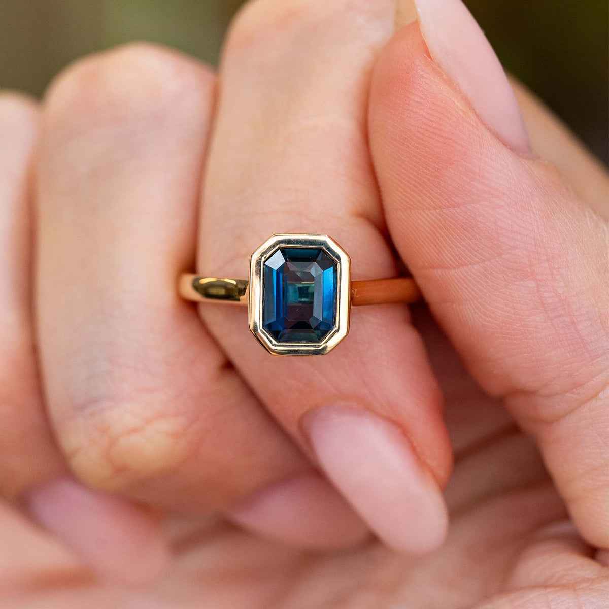 A close-up of hand holding an Antique-inspired, Deep blue sapphire solitaire engagement ring with a yellow gold bezel setting and wedding fit band. 