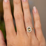 A close-up of a hand wearing a yellow gold Antique-inspired engagement ring with a central oval green sapphire surrounded by a halo of round-cut diamonds. 