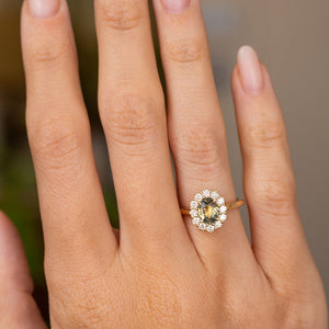 A close-up of a hand wearing a yellow gold Antique-inspired engagement ring with a central oval green sapphire surrounded by a halo of round-cut diamonds. 