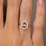 A close-up of a hand wearing a yellow gold halo ring with a central oval green sapphire and diamond accent stones. Antique-inspired with a yellow gold band. 
