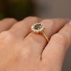 A close-up of a yellow gold engagement ring featuring a cluster of diamonds and  a central oval green Sri Lankan sapphire, worn on a person's finger.  Showcasing the antique-inspired design and wedding fit of the band. 