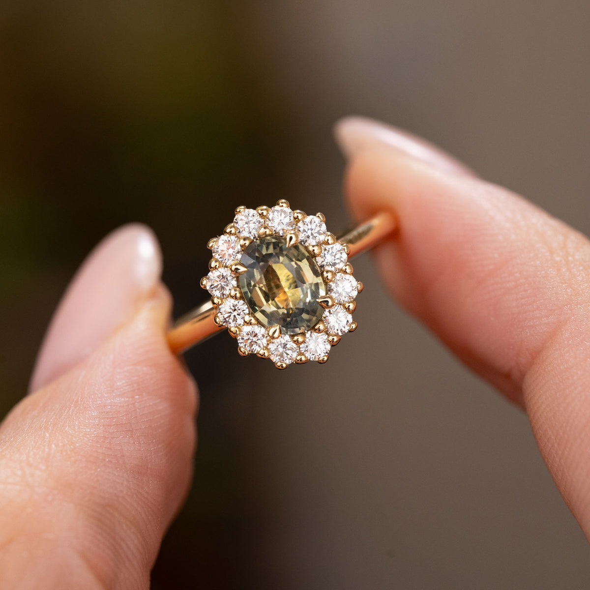 A close-up of the central oval green Sri Lankan sapphire and surrounding halo of diamonds on an Antique inspired cluster engagement ring.  The ring is held between two fingers by the yellow gold band. 