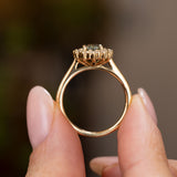A close-up of an Antique inspired green sapphire and diamond cluster engagement ring, showcasing the side profile, wedding fit and yellow gold band.  Held between two fingers against a blurred brown and green background.