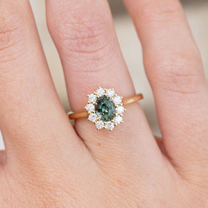 Detailed hand close-up highlighting the sapphire’s varied green tones and the floral-style halo of round brilliant diamonds.