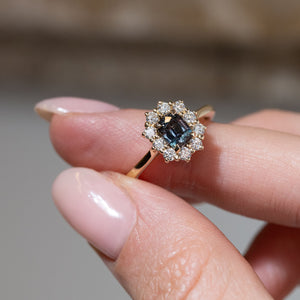 Hand-held close-up of the dark blue sapphire surrounded by sparkling white diamonds in an antique-inspired yellow gold setting.