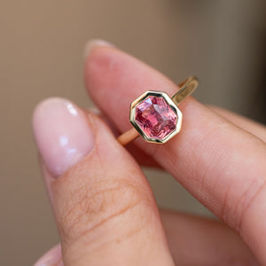 Antique-style pink sapphire solitaire held between the fingers, offering a detailed look at its facets and rub-over frame.