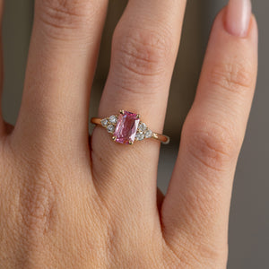 Close hand detail highlighting the soft pink sapphire, three-diamond shoulder clusters and polished yellow gold band.