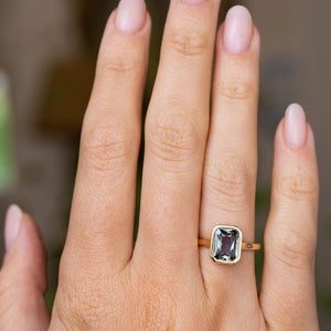 A close-up of a hand wearing an Antique-inspired, Charcoal-blue sapphire solitaire engagement ring in yellow gold. The ring is shown on the ring finger against a softly blurred, neutral background.