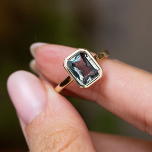 A yellow gold engagement ring with a rectangular emerald-cut blue grey sapphire held between fingers against a blurred green and brown background. A solitaire ring inspired by Antique designs.