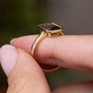 A close-up of a Vintage-inspired, yellow gold solitaire engagement ring.  Displaying the low profile, gold band and bezel set blue sapphire, being held between fingers.
