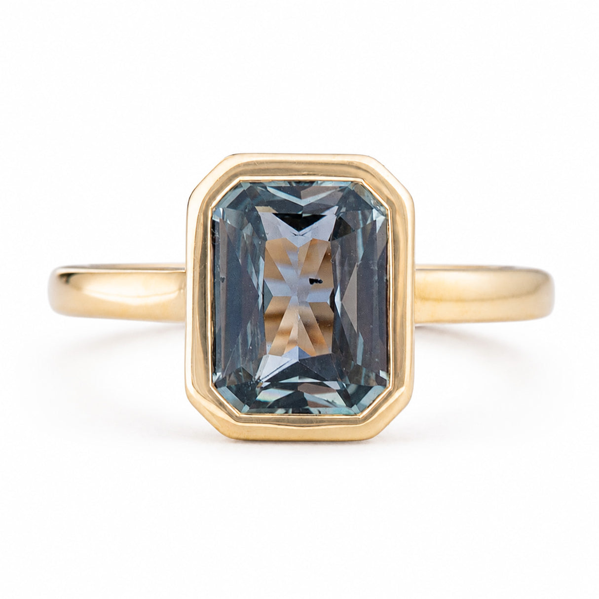 An Antique-inspired, yellow gold solitaire engagement ring featuring a large, faceted emerald-cut blue-gray sapphire in an octagonal bezel setting, shown on a clean white background. 